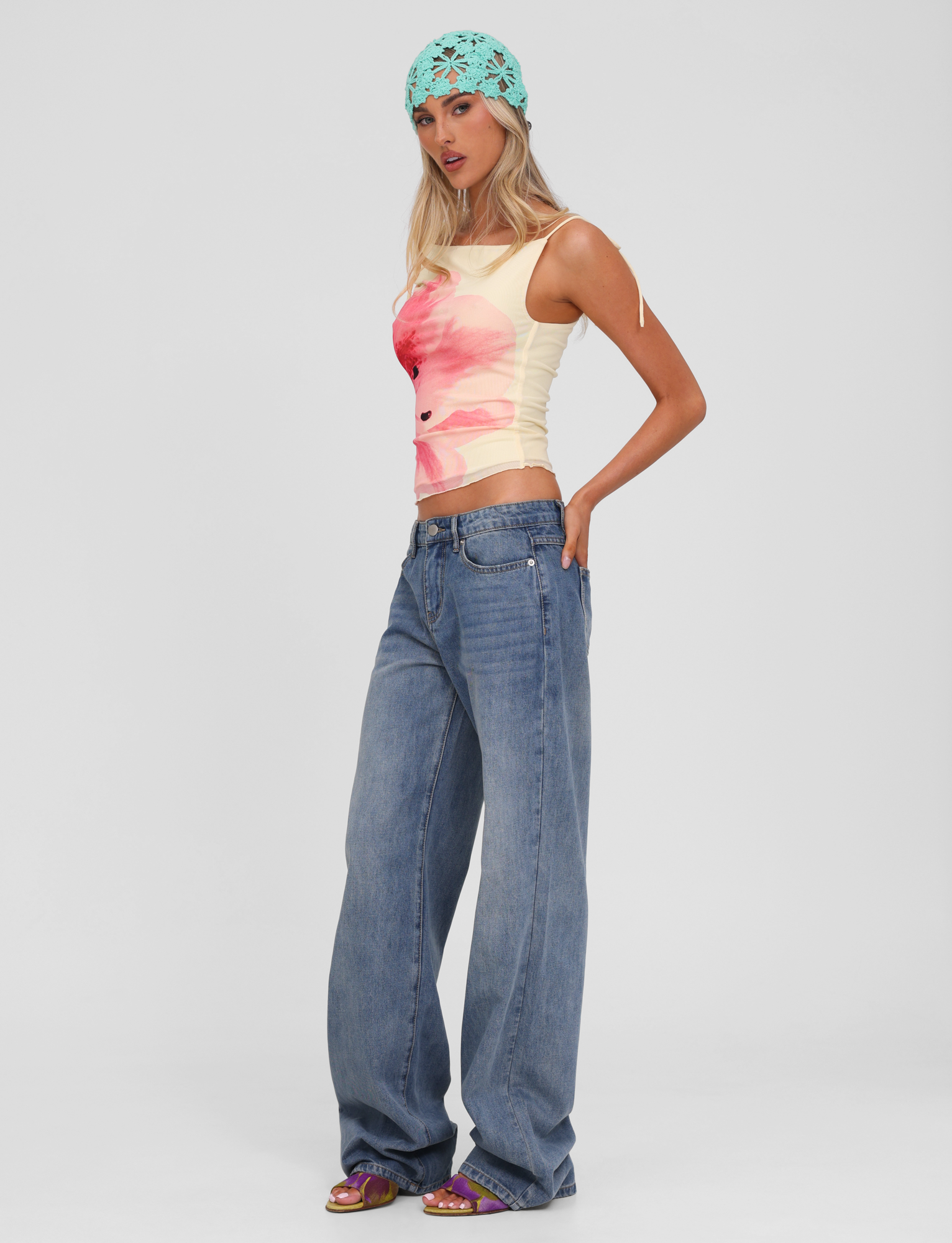 A woman poses against a plain light background wearing a light yellow floral tank, the TIGERMIST ESTRELLA JEAN - MID WASH with a button fly, heeled sandals, and a light blue crocheted headscarf.