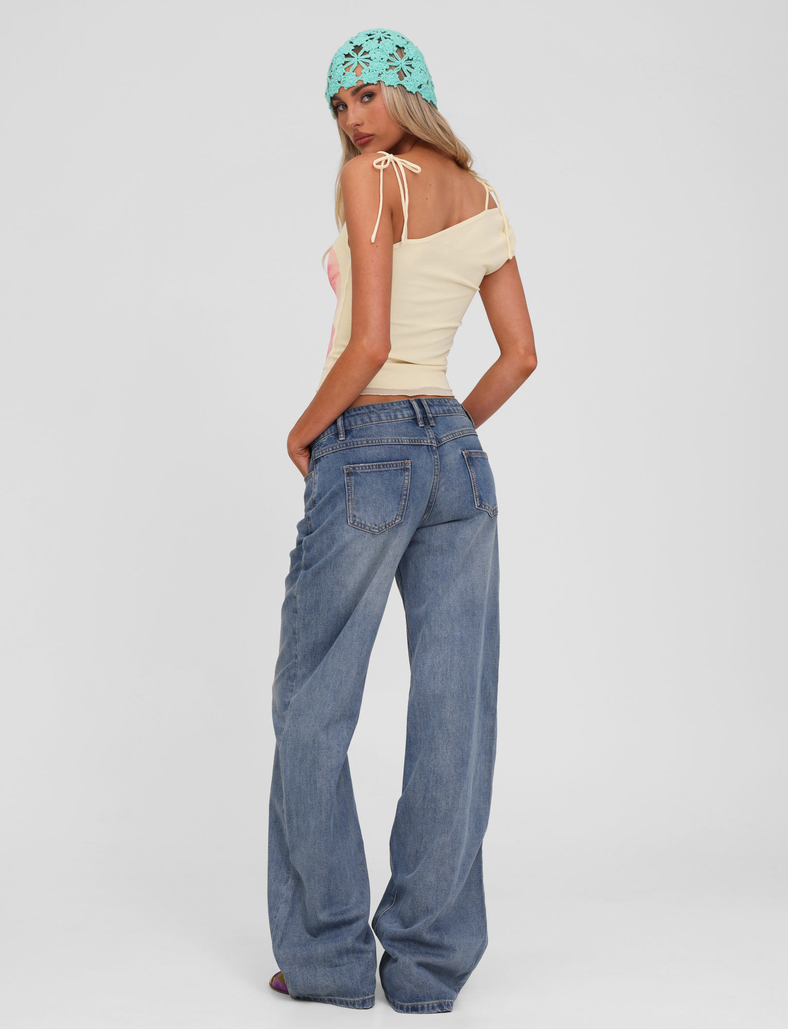 A woman in a light yellow top, TIGERMIST ESTRELLA JEAN - MID WASH straight leg jeans with a button fly, and a light blue crochet headscarf stands facing away, glancing over her shoulder against a plain white background.