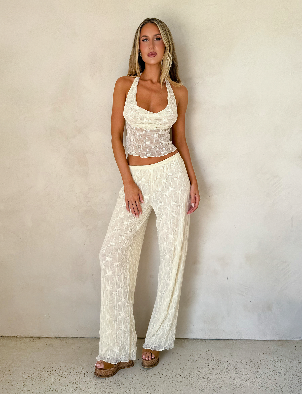 A woman with long, straight blonde hair stands against a plain wall wearing TIGERMIST's KATERINA PANT in ivory lace, featuring floral lace details and an elasticated waist, paired with a sheer, cream halter top and platform sandals.