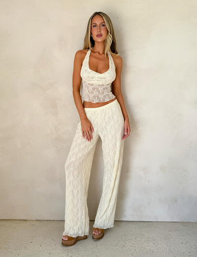 A woman with long, straight blonde hair stands against a plain wall wearing TIGERMIST's KATERINA PANT in ivory lace, featuring floral lace details and an elasticated waist, paired with a sheer, cream halter top and platform sandals.