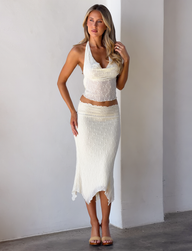 A woman with long, wavy blonde hair wears the TIGERMIST KATERINA MIDI SKIRT in cream and a white halter top, standing indoors against a white wall and floor, looking slightly to the side.