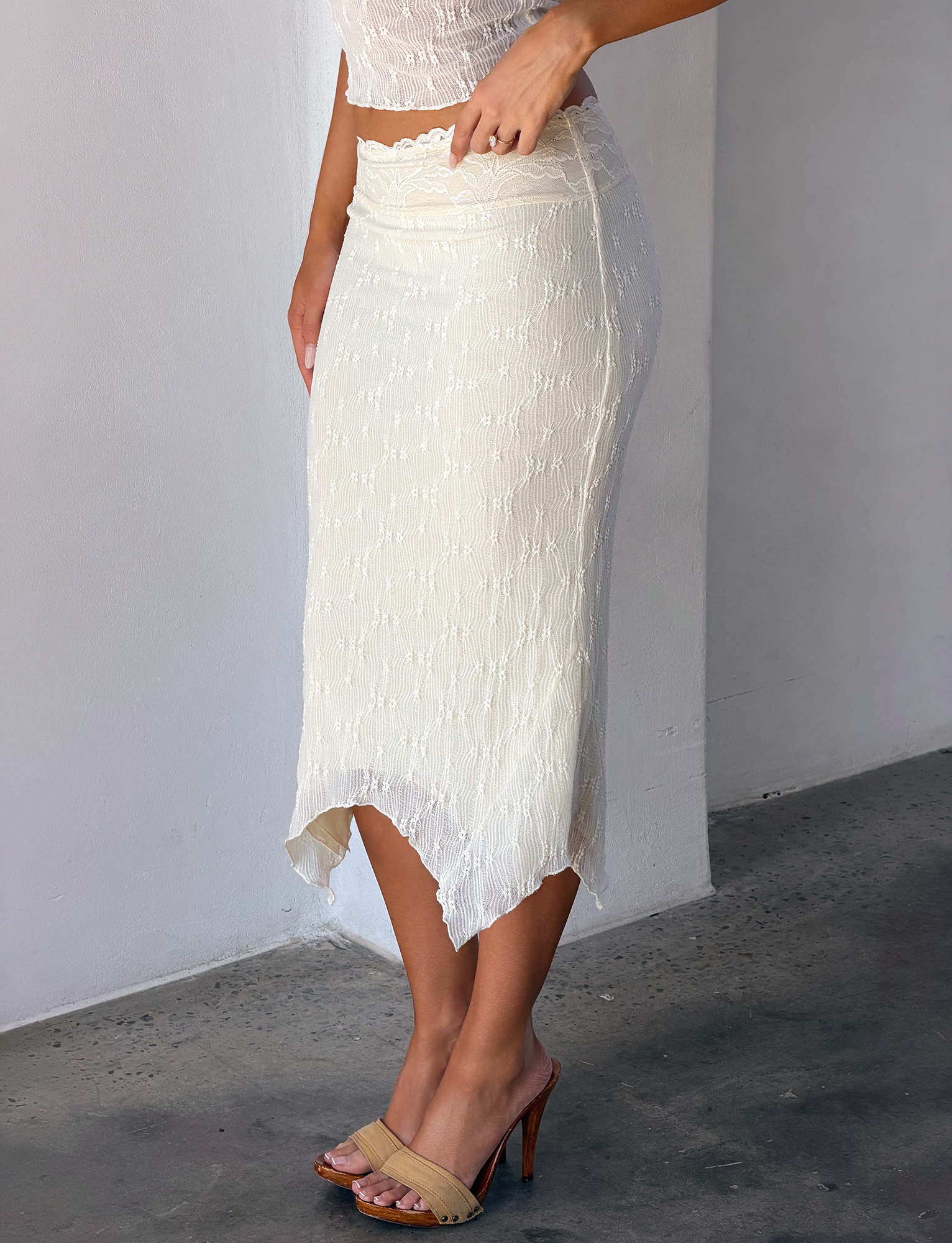 A woman wearing the TIGERMIST KATERINA MIDI SKIRT in cream, featuring an asymmetrical hem, paired with a coordinating white top and tan heels, stands on a concrete floor by a light-colored wall.