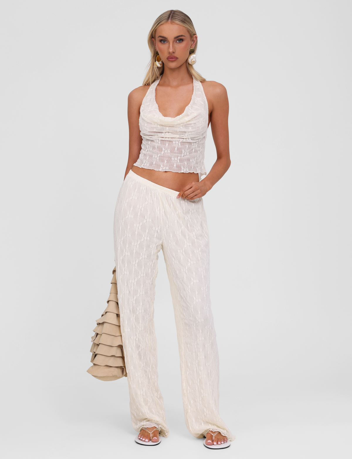 A woman poses against a plain background wearing the TIGERMIST KATERINA TOP in ivory lace with matching pants. She carries a beige ruffled handbag, wears white sandals, and her hair is styled in loose waves.