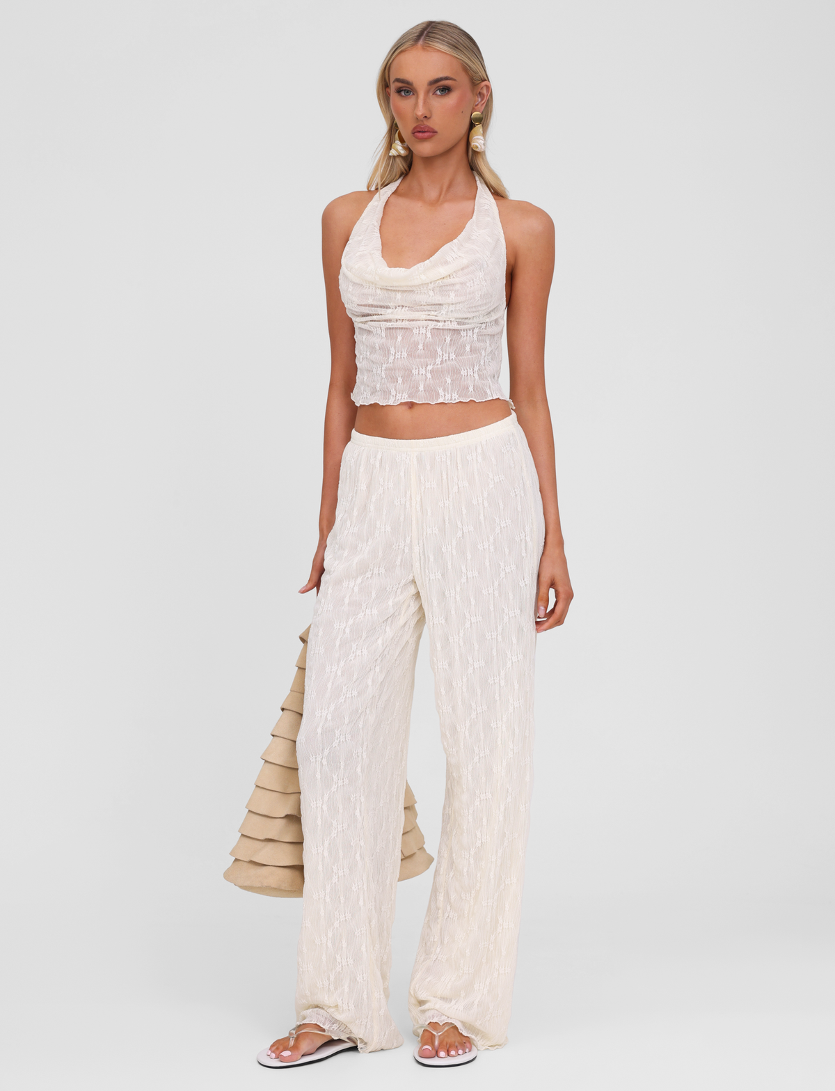 A woman stands against a white background wearing TIGERMIST’s cream-colored sleeveless halter top and KATERINA PANT in ivory lace, paired with sandals and a beige ruffled bag.