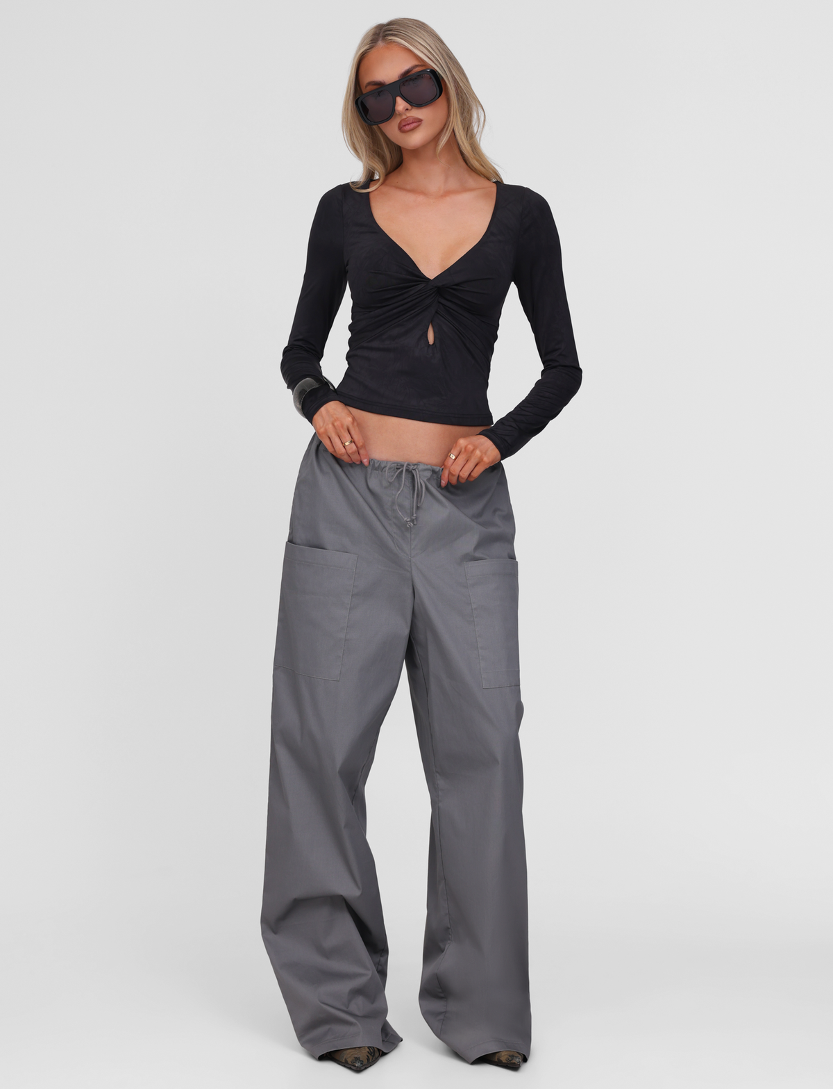 A woman wearing oversized sunglasses, loose gray pants, and the TIGERMIST SAFFIRA LONG SLEEVE TOP in black stands against a plain white background.