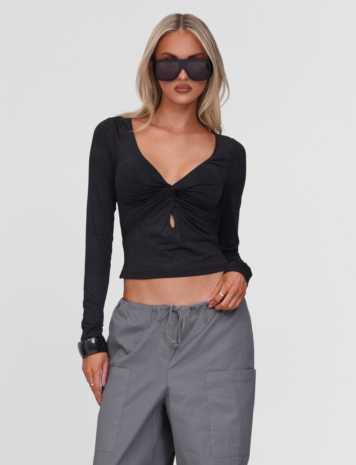A woman with long blonde hair wears oversized black sunglasses, gray drawstring pants with pockets, and the TIGERMIST SAFFIRA LONG SLEEVE TOP in black, standing against a plain white background.