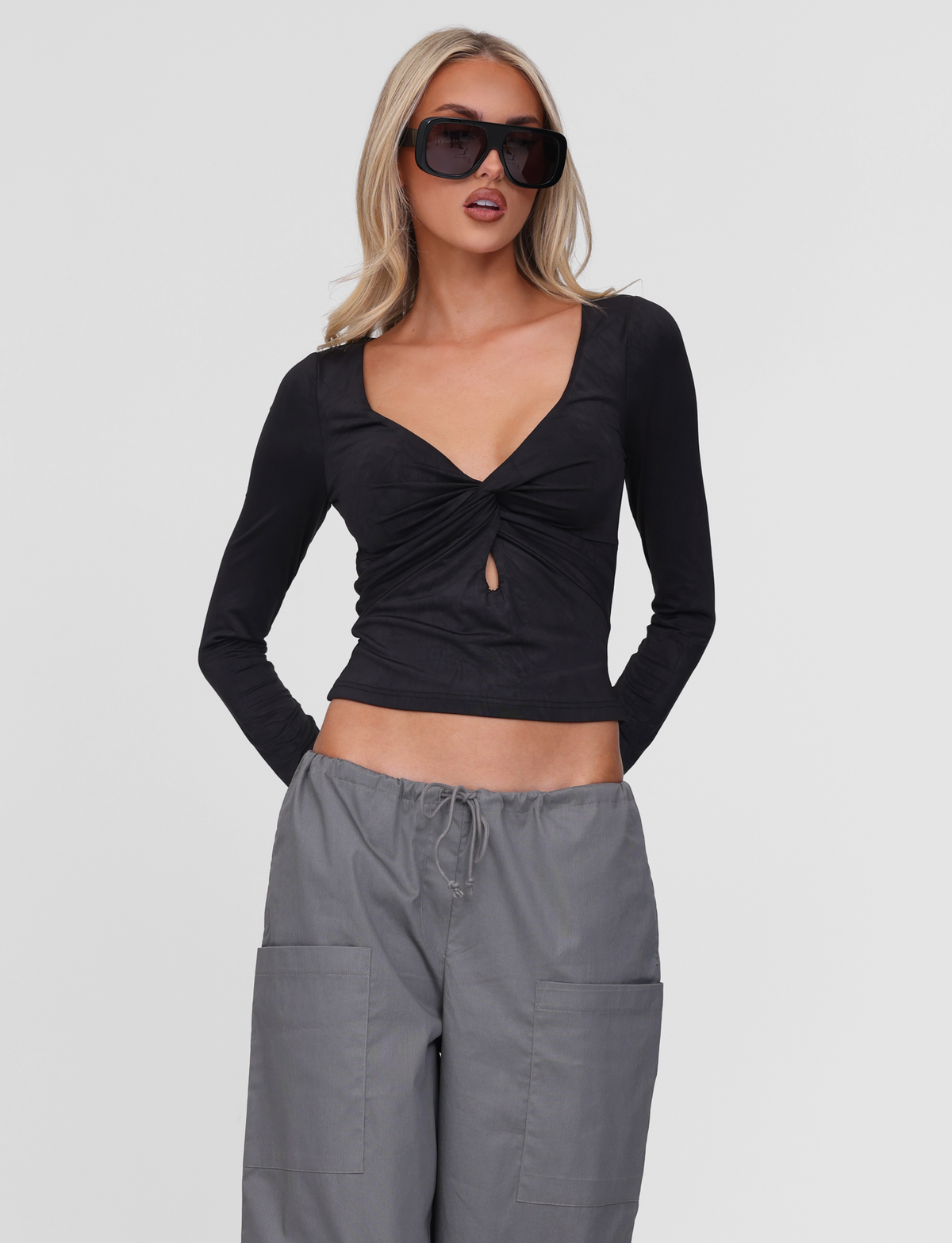 A woman with long blonde hair wears large black sunglasses and the TIGERMIST SAFFIRA LONG SLEEVE TOP in black, featuring a knot and keyhole chest detail, paired with loose gray drawstring pants. She stands against a plain white background.