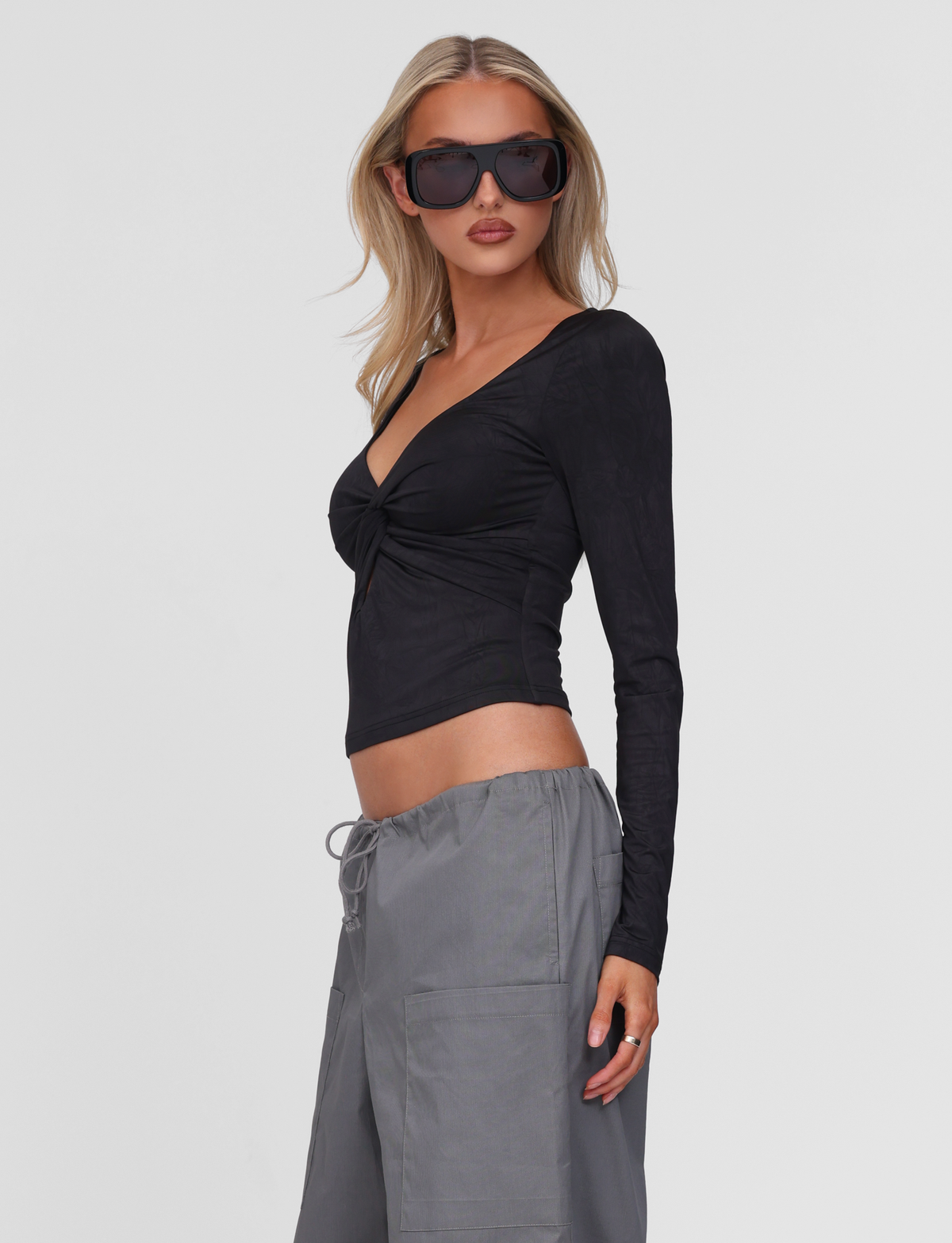A woman with blonde hair wears large dark sunglasses, loose gray drawstring pants, and the TIGERMIST SAFFIRA LONG SLEEVE TOP in black, featuring a low-cut V neckline, standing against a plain white background.