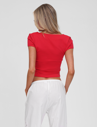 A person with long blonde hair, seen from the back against a plain light background, wears the TIGERMIST SAVINA TOP in red—fitted with embroidered button details—and loose white pants.