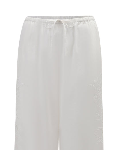 The CRESSY PANT - WHITE by TIGERMIST features a wide-leg, full-length cotton design with an elastic waistband and drawstring, soft lightweight fabric, and subtle floral embroidery along the sides.