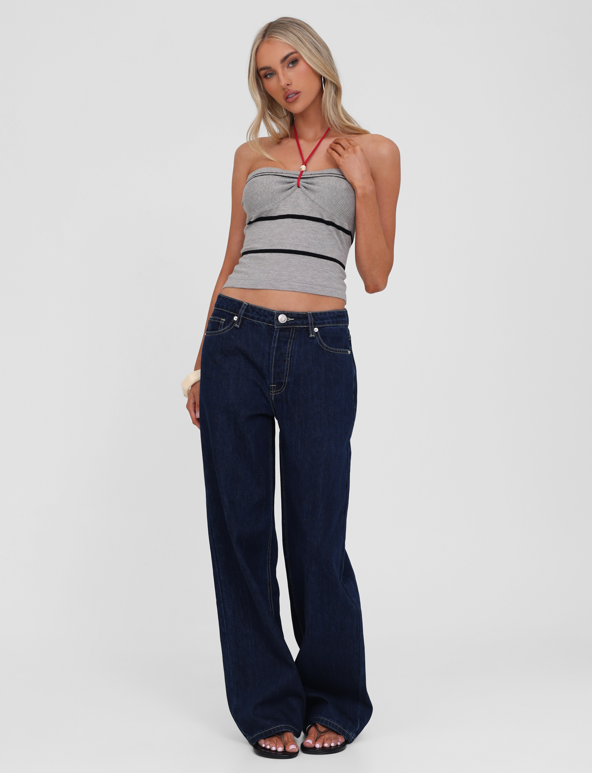 A woman with long blonde hair poses casually against a plain background, wearing the TIGERMIST NOUR TOP in grey stripe, dark blue wide-leg jeans, and open-toed shoes.
