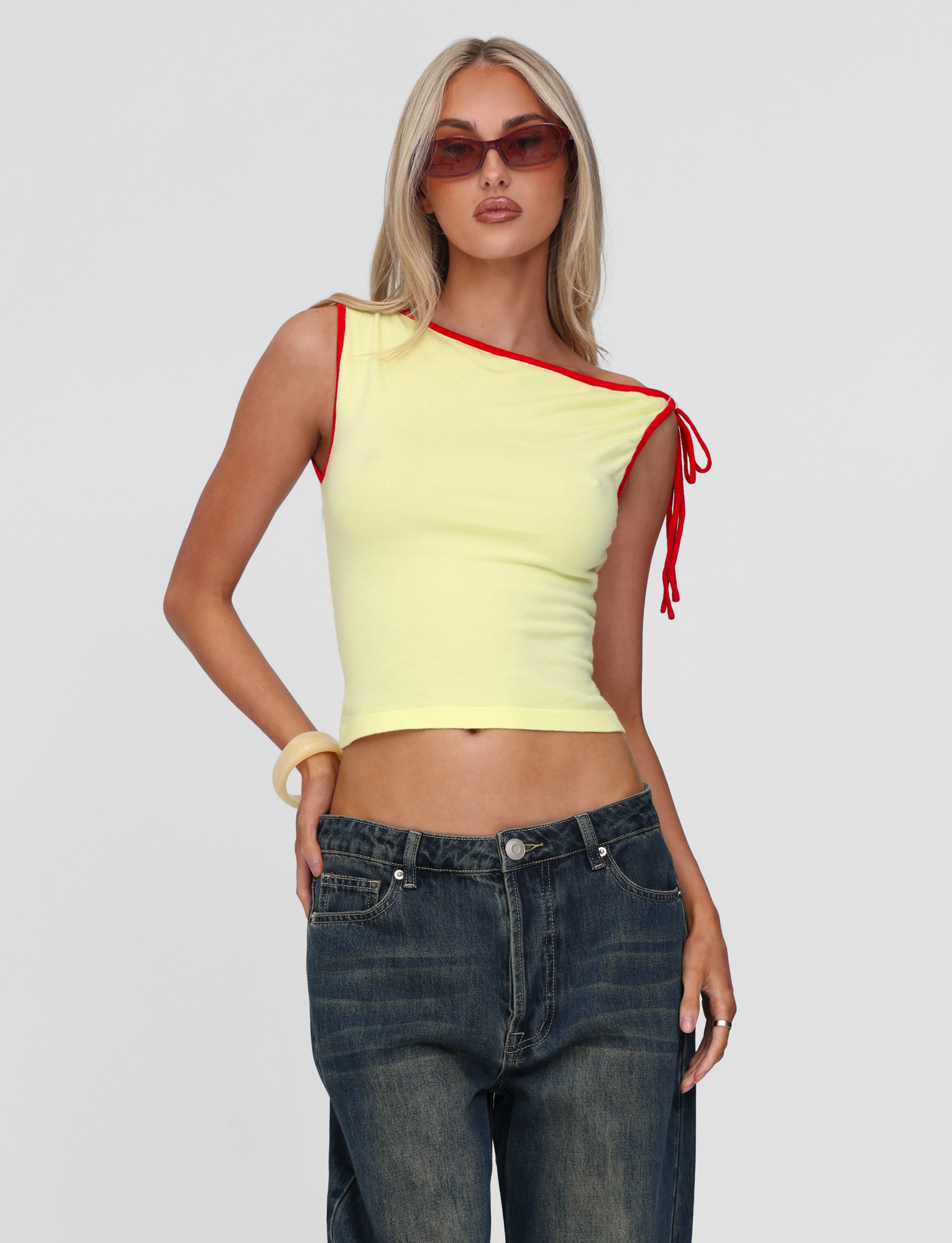 A woman with long blonde hair poses against a white background in the TIGERMIST ALIRA TOP - YELLOW featuring a red contrast neckline, styled with red sunglasses, blue jeans, and a chunky bracelet.