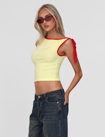A woman with long blonde hair and sunglasses wears high-waisted blue jeans and the TIGERMIST ALIRA TOP in yellow, featuring a red contrast neckline and shoulder tie, standing against a plain light gray background.