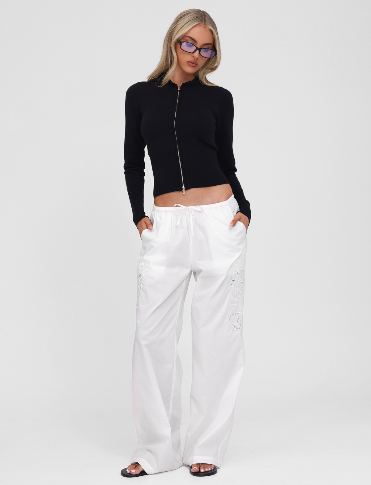A person stands against a plain white background with hands in pockets, wearing the SARINA JUMPER - BLACK by TIGERMIST, paired with loose white drawstring pants, black sandals, and blue-tinted glasses.