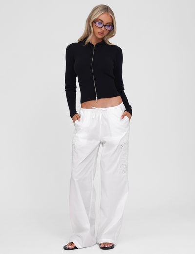 A person stands against a plain white background with hands in pockets, wearing the SARINA JUMPER - BLACK by TIGERMIST, paired with loose white drawstring pants, black sandals, and blue-tinted glasses.