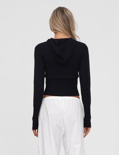 A person with long, wavy blonde hair stands with their back to the camera, wearing the TIGERMIST SARINA JUMPER in black and loose white pants against a plain light background.