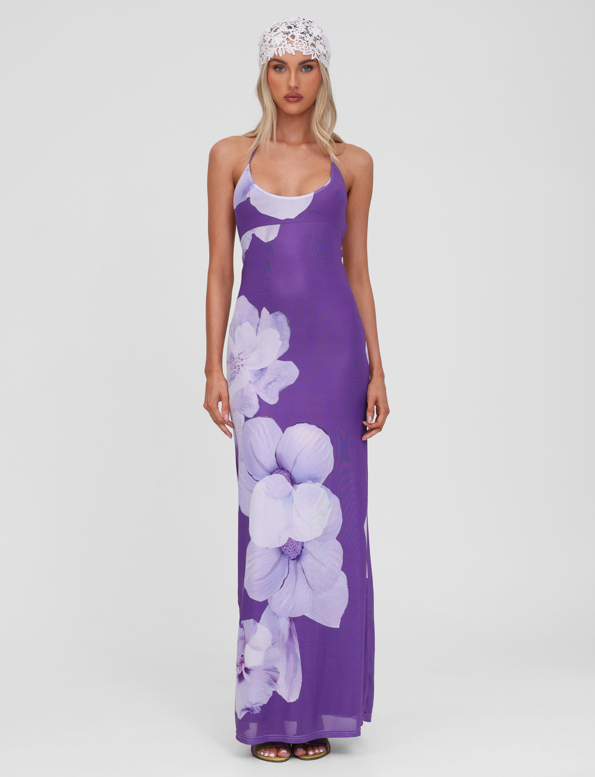 A woman faces forward wearing the TIGERMIST NOUR MESH MAXI DRESS in Purple Macro Floral, styled with a white lace headscarf and brown sandals. The sleeveless dress features a plunging back. The background is plain white.