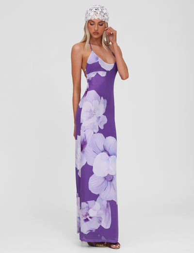 A woman models the TIGERMIST NOUR MESH MAXI DRESS in purple macro floral with a white lace headscarf, standing against a white background, one hand to her face and gazing downward thoughtfully.