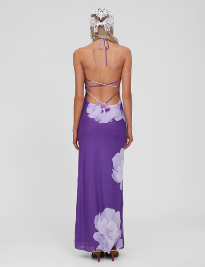 A woman stands facing away, wearing the TIGERMIST NOUR MESH MAXI DRESS in purple macro floral with crisscross straps and a white lace headpiece against a plain light gray background.