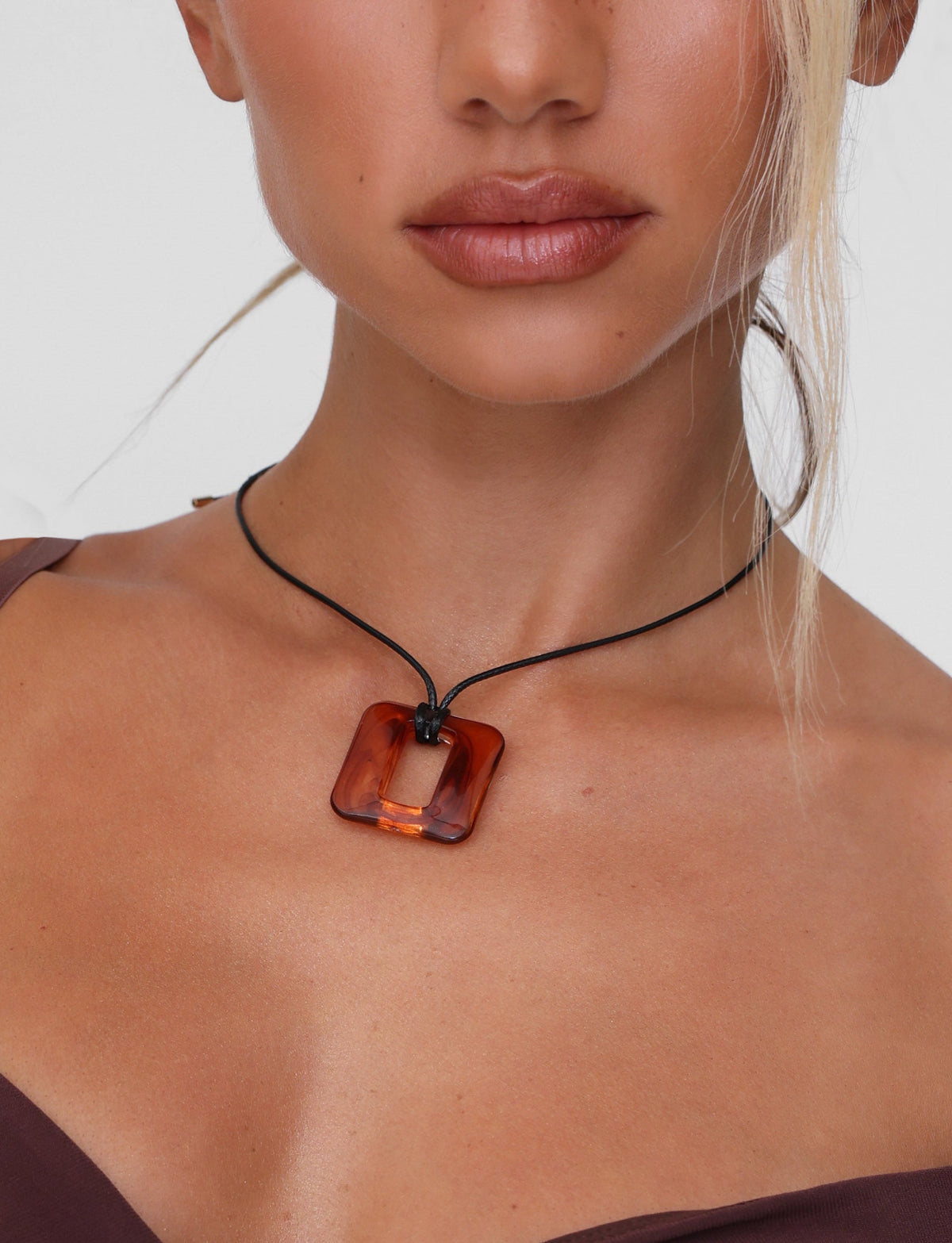 A close-up shows a person wearing the MIRREN SQUARE NECKLACE - BROWN by TIGERMIST, featuring a black woven cord and square amber pendant with resin. The minimalist design stands against a plain white background, with their hair partially visible and falling loosely.