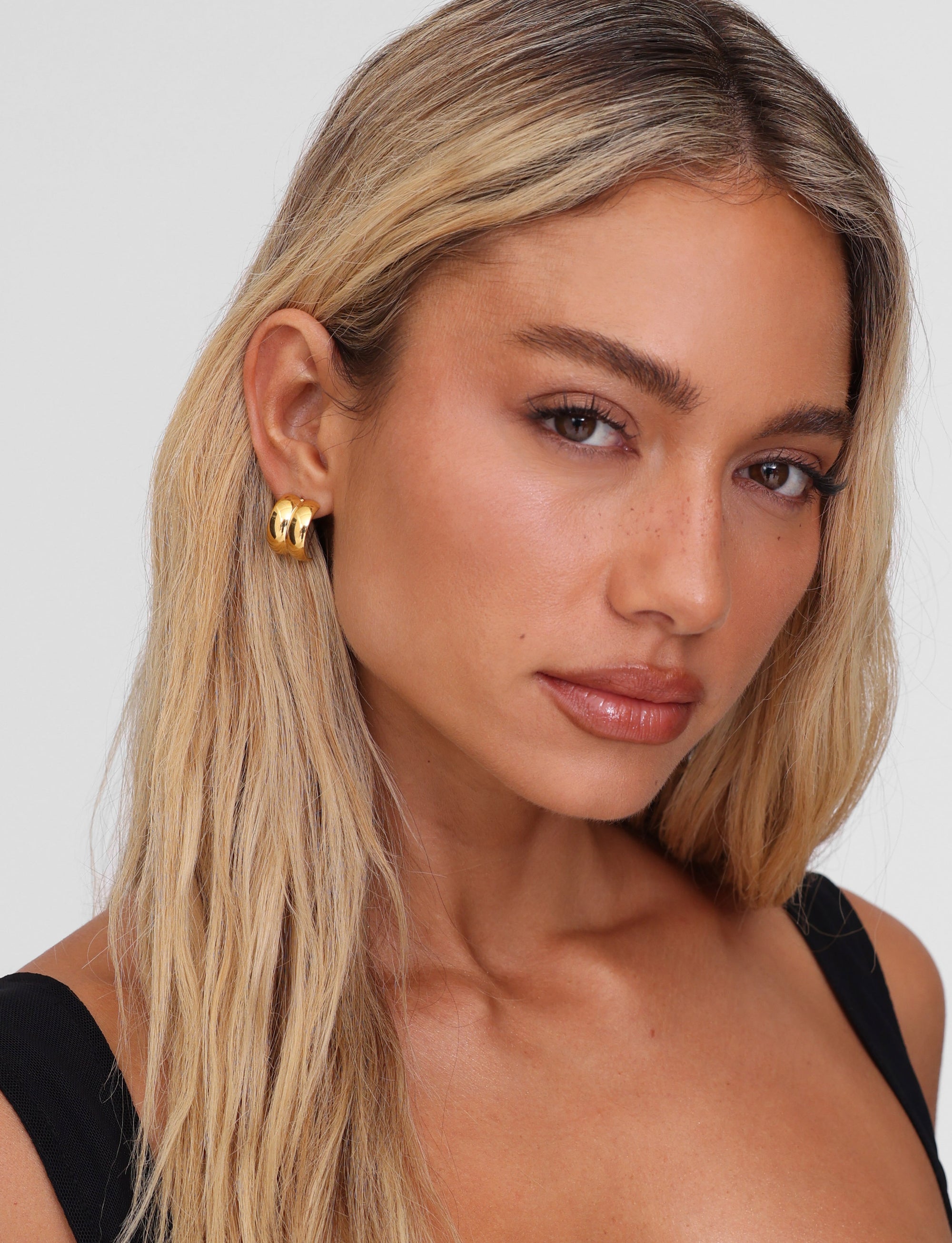 A woman with long blonde hair and neutral makeup looks at the camera, wearing a black top and bold LILLIA EARRING - GOLD by TIGERMIST. The background is plain white.