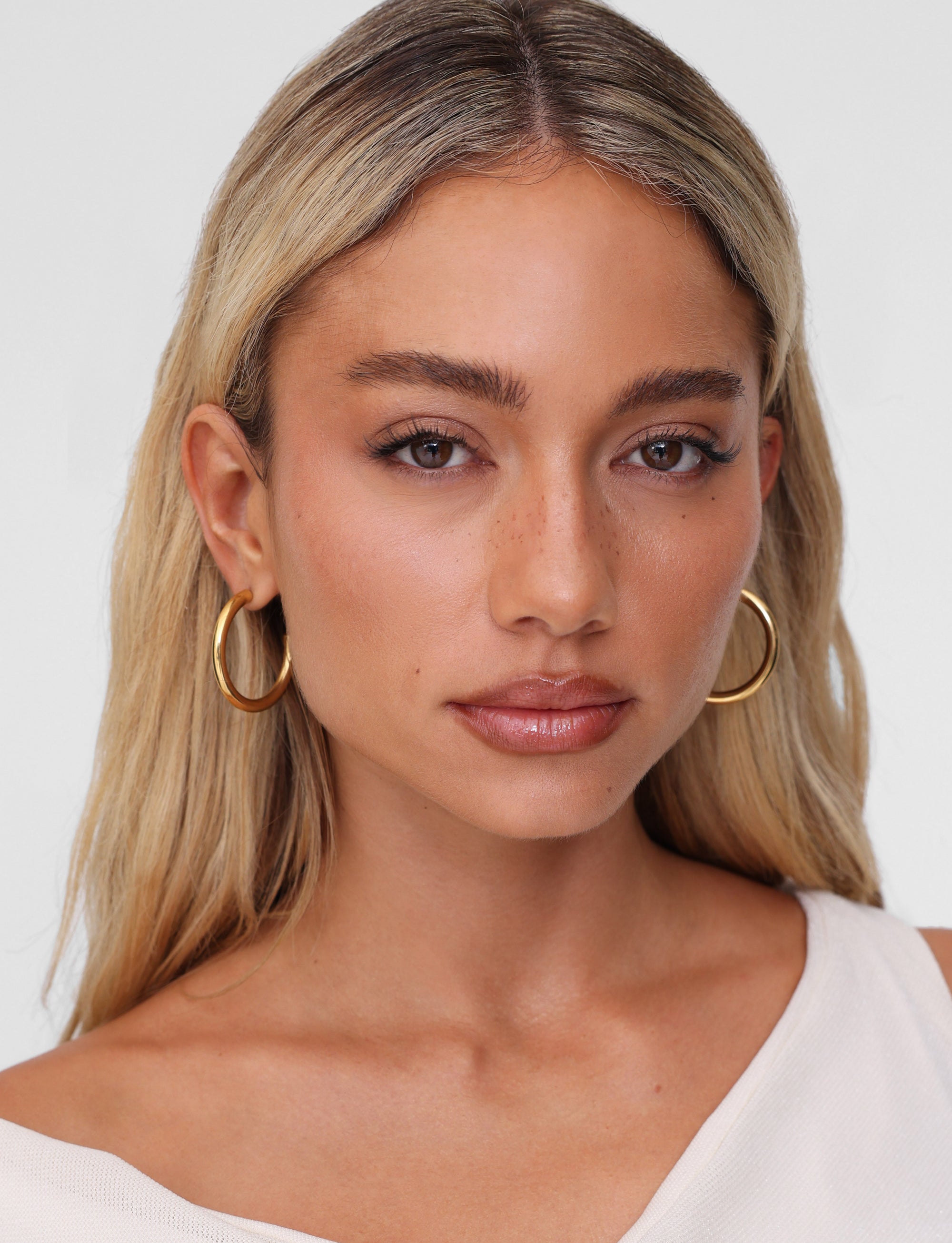 A woman with long blonde hair wears TIGERMIST's PIPER EARRING - GOLD, paired with a cream top against a neutral backdrop. Her serene expression and natural makeup accentuate the elegance of these hypoallergenic accessories.