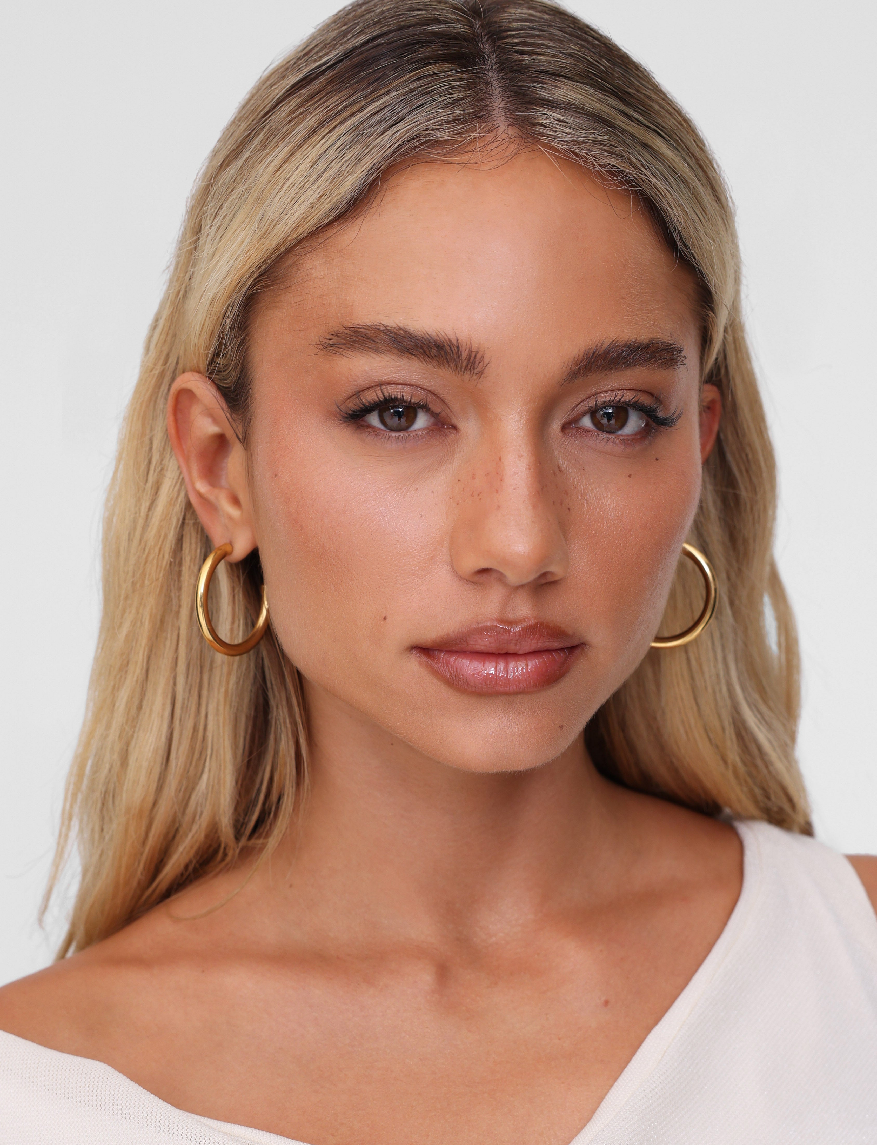 A woman with long blonde hair wears TIGERMIST's PIPER EARRING - GOLD, paired with a cream top against a neutral backdrop. Her serene expression and natural makeup accentuate the elegance of these hypoallergenic accessories.