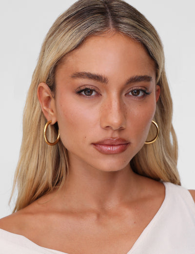 A woman with long blonde hair wears TIGERMIST's PIPER EARRING - GOLD, paired with a cream top against a neutral backdrop. Her serene expression and natural makeup accentuate the elegance of these hypoallergenic accessories.