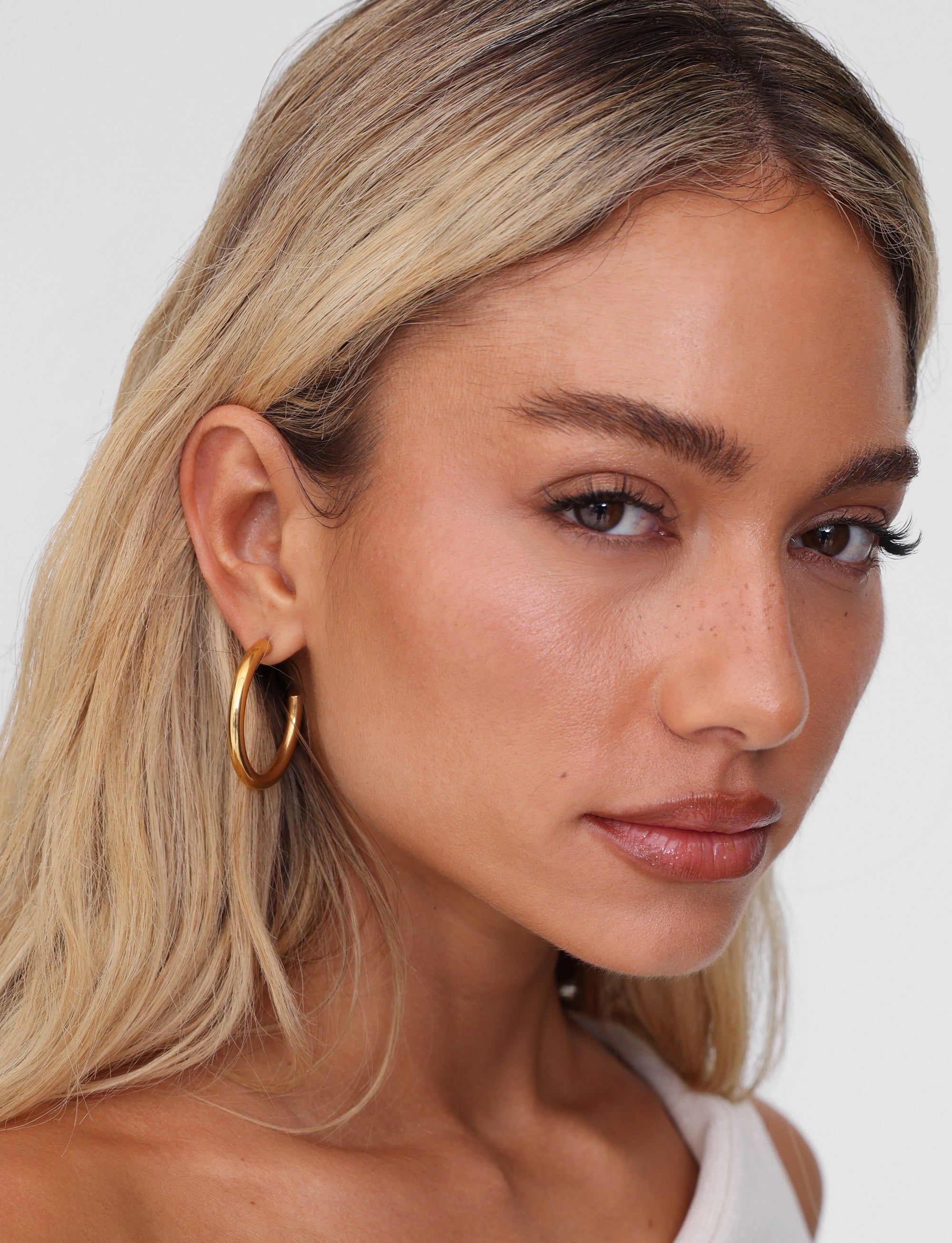 A person with long blonde hair is gazing into the camera with a neutral expression, wearing TIGERMIST's PIPER EARRING - GOLD. They are dressed in a pale top against a plain white background.
