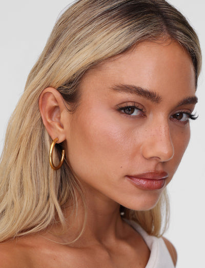 A person with long blonde hair is gazing into the camera with a neutral expression, wearing TIGERMIST's PIPER EARRING - GOLD. They are dressed in a pale top against a plain white background.