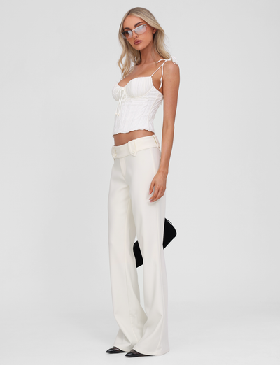 A woman with long blonde hair and glasses stands against a white background wearing the TIGERMIST RANIA TOP in off-white, featuring strappy shoulders and a sweetheart neckline, paired with high-waisted wide-leg white pants and holding a black clutch.