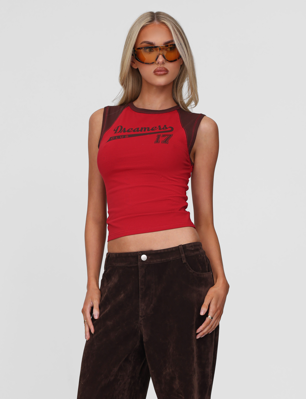 A woman showcases relaxed Dreamers Club style in TIGERMIST’s FIERA TEE - RED with “Dreamers 17” print, brown pants, and oversized brown sunglasses, standing confidently against a plain white background.