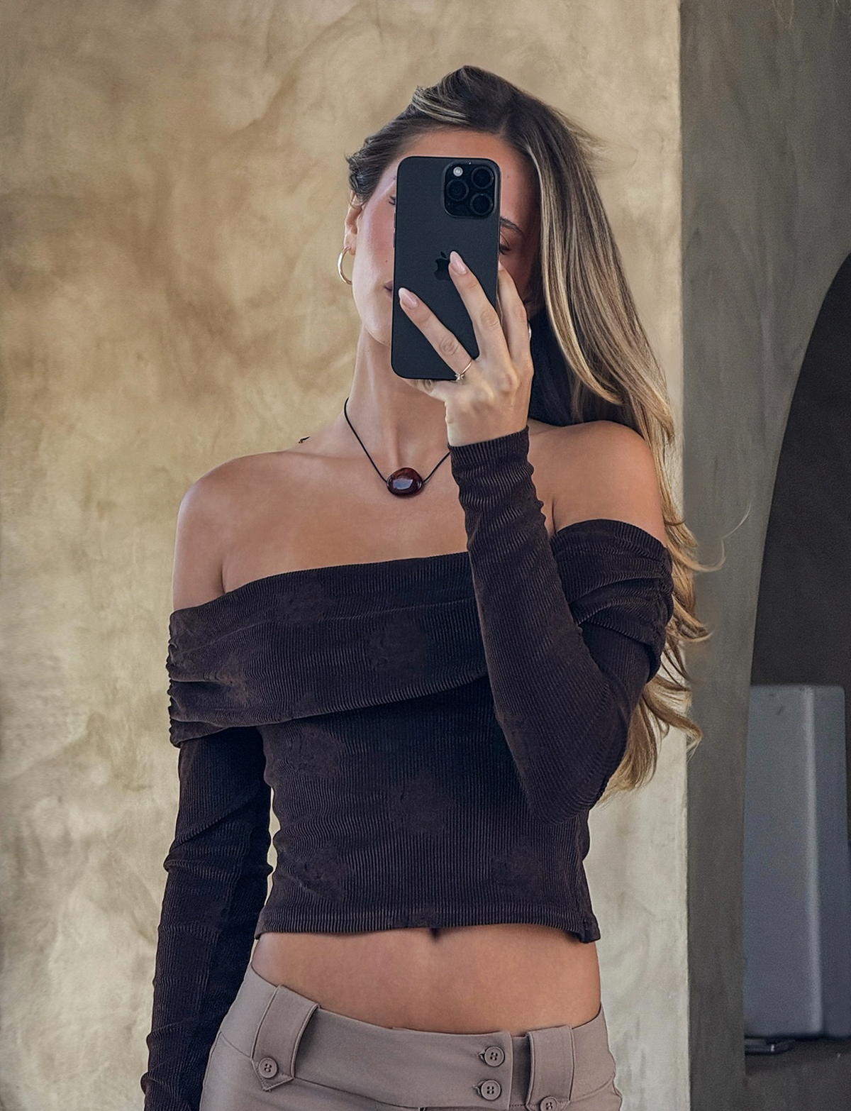 A woman with long hair takes a mirror selfie, holding her phone in front of her face. She wears the TIGERMIST KORRA TOP in Chocolate Rose, paired with light beige pants and a round pendant necklace. The background is neutral.
