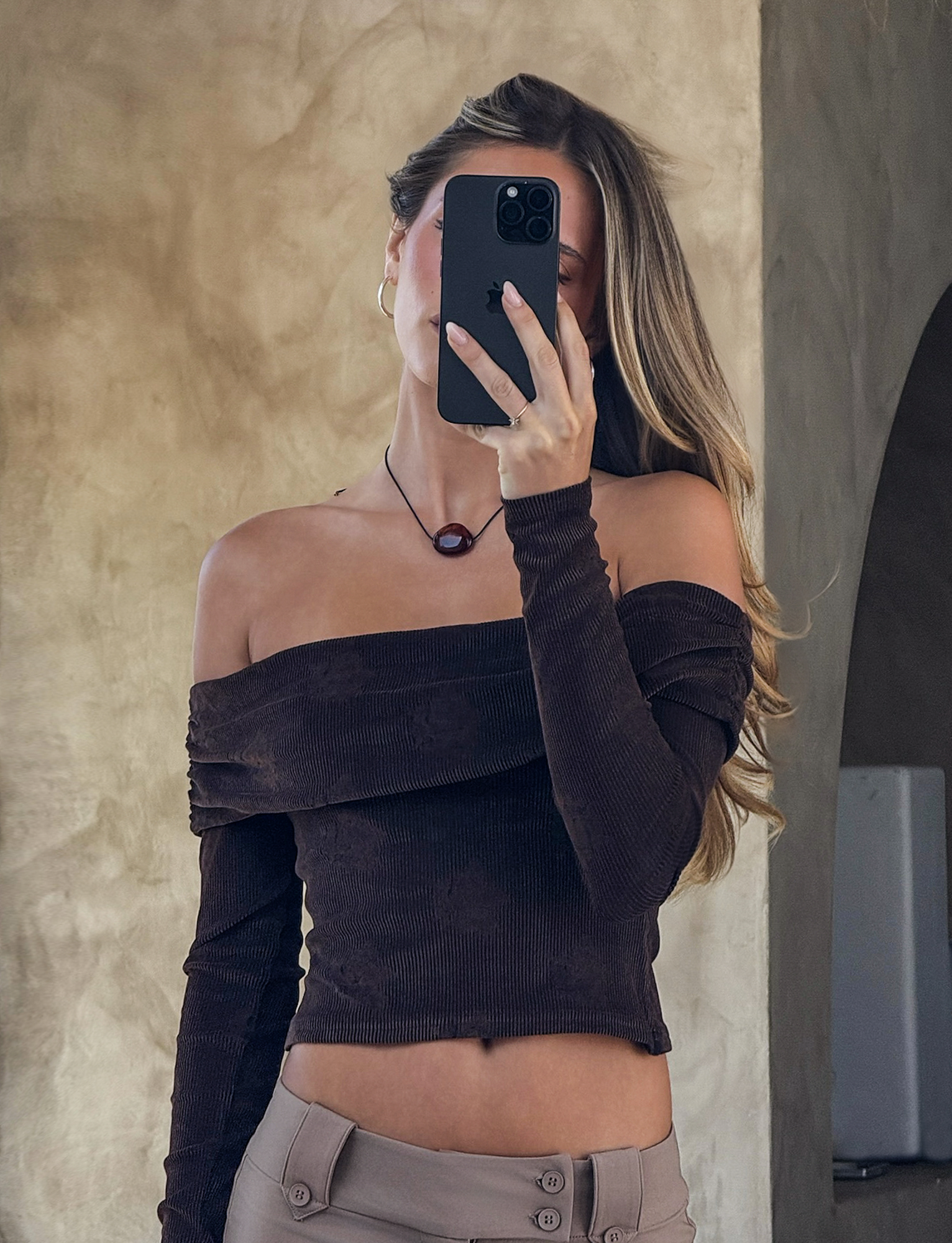 A woman with long hair takes a mirror selfie, holding her phone in front of her face. She wears the TIGERMIST KORRA TOP in Chocolate Rose, paired with light beige pants and a round pendant necklace. The background is neutral.