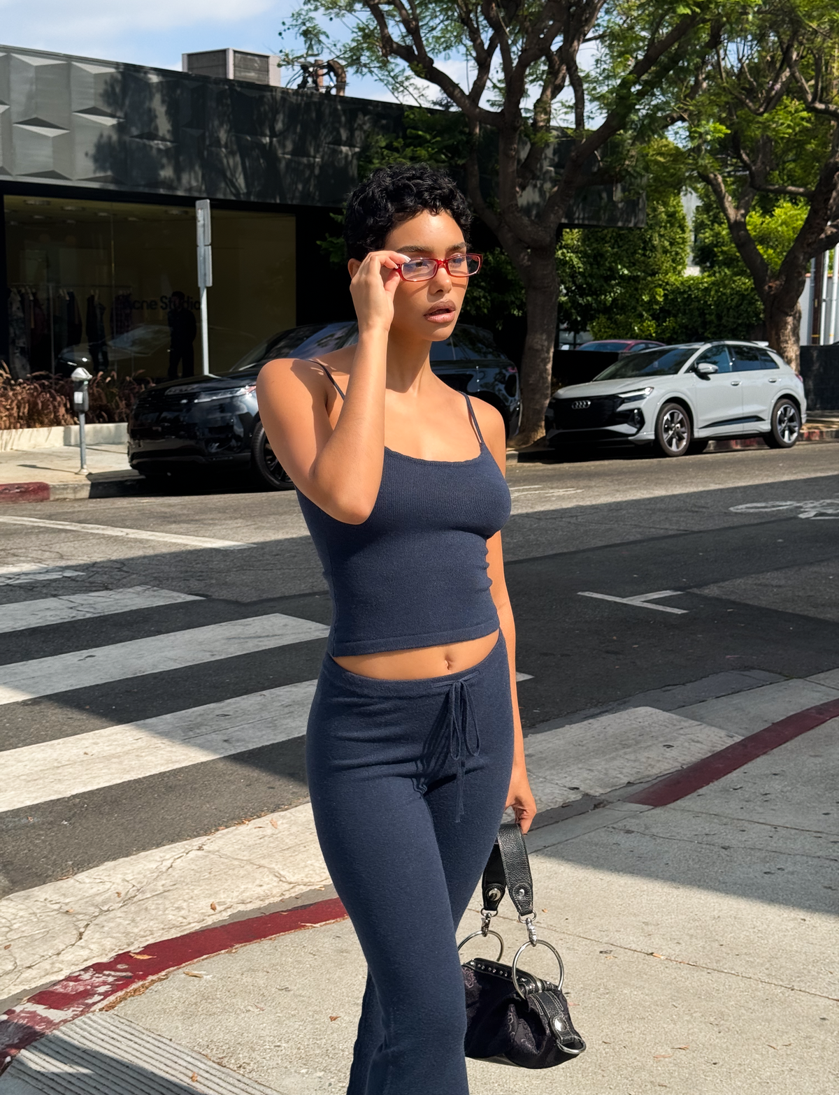 Wearing the TIGERMIST LEORA TOP - NAVY with adjustable straps and matching pants, a person stands on a city sidewalk holding a black handbag and adjusting red sunglasses. Cars, trees, and a crosswalk are in the background.