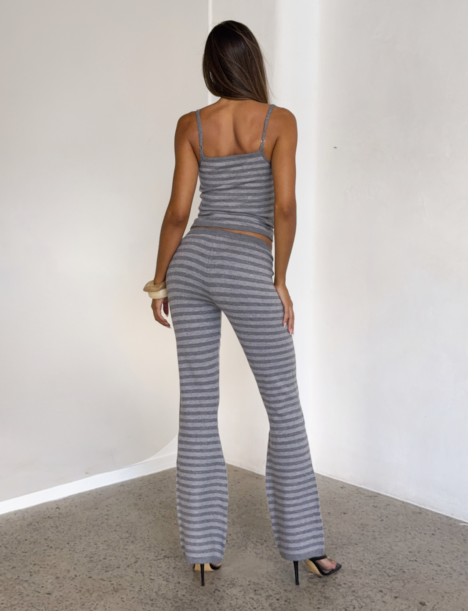 A woman with long brown hair, facing away indoors, wears the TIGERMIST LEORA PANT - GREY STRIPE with a matching striped tank top and black heels, one hand on her hip against a minimalistic background.