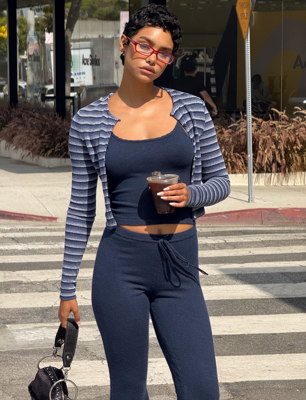 A woman with short hair and red glasses stands on a city crosswalk wearing the TIGERMIST TIMARA CARDIGAN - NAVY STRIP, matching pants, and a navy blue knit crop top, holding an iced coffee and a small handbag.