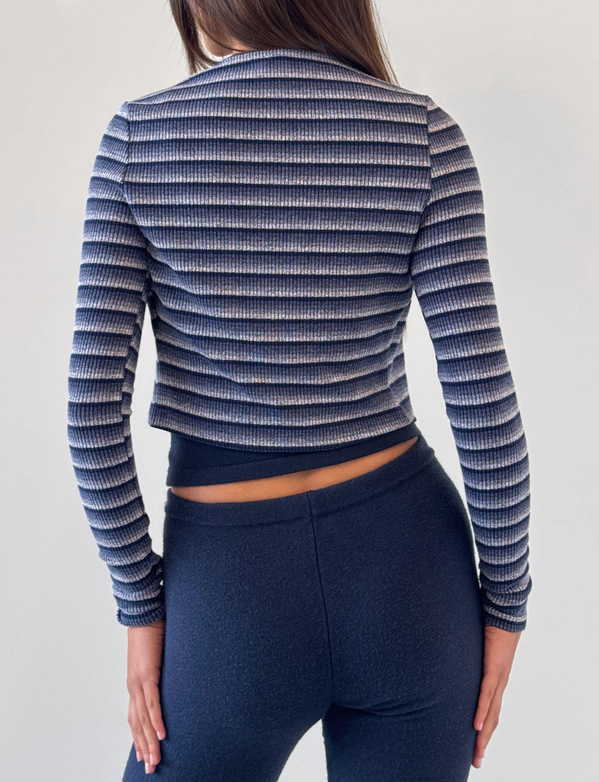A woman faces away against a plain background, wearing TIGERMIST's LEORA PANT in navy—a dark, relaxed knit pant with a drawstring waist—paired with a blue and white long-sleeve striped top. Her right arm rests by her side.