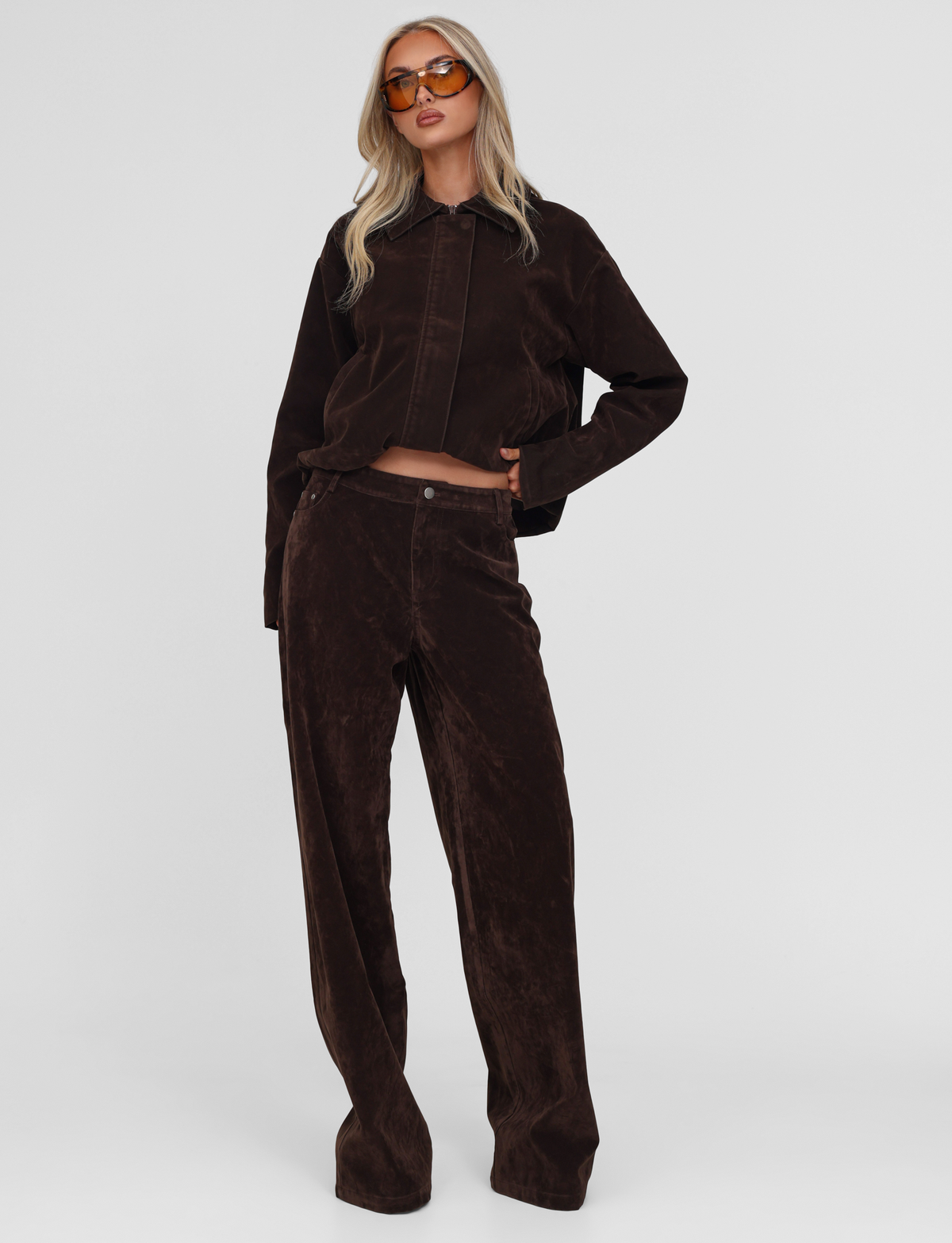 A woman with long blonde hair confidently poses against a white background, wearing orange sunglasses, a loose dark brown jacket, and TIGERMIST’s CAMELLIA PANT in chocolate suede.