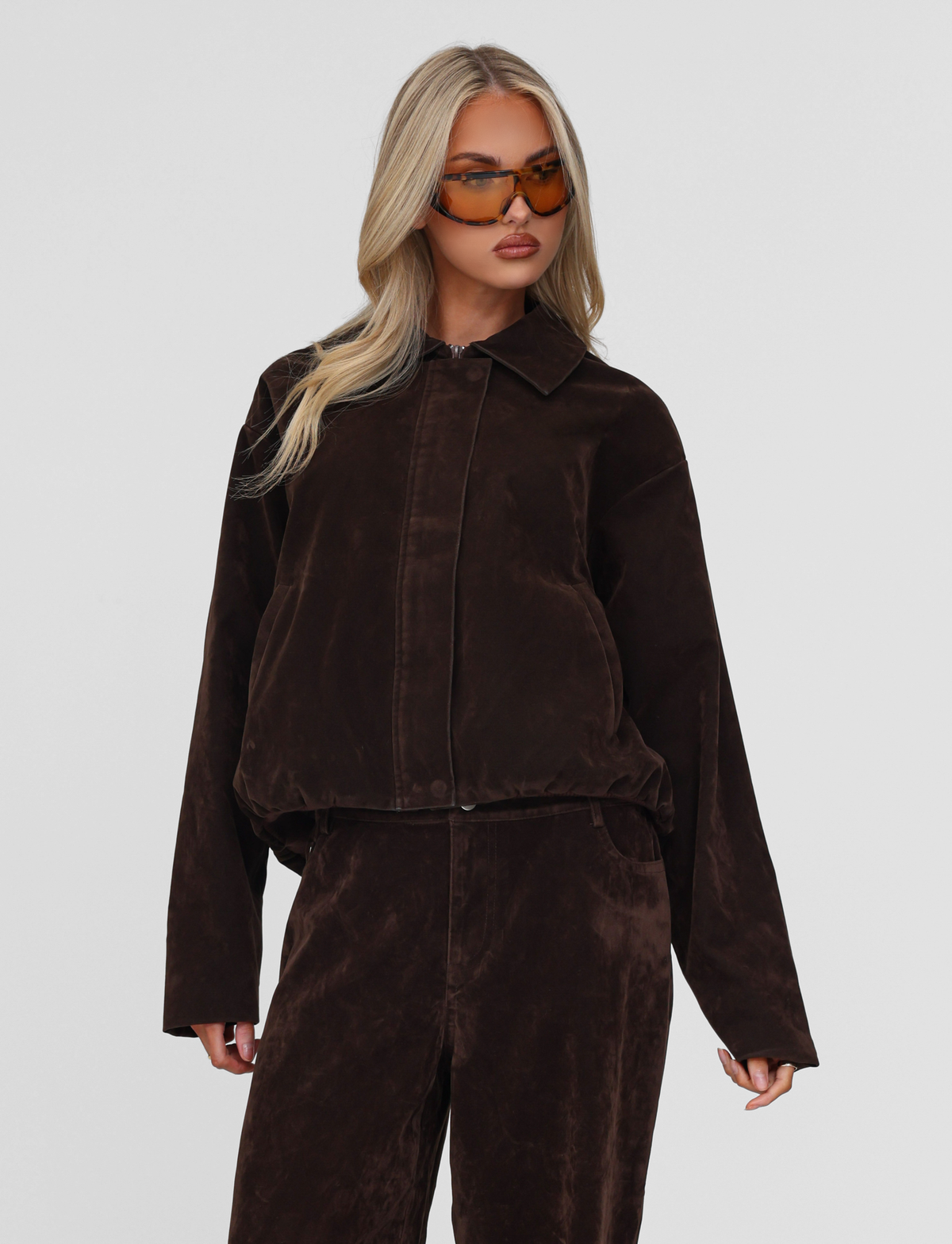 A woman with long blonde hair wears large brown sunglasses and the TIGERMIST CAMELLIA BOMBER JACKET - CHOCOLATE SUEDE with matching pants, standing against a plain light gray background.