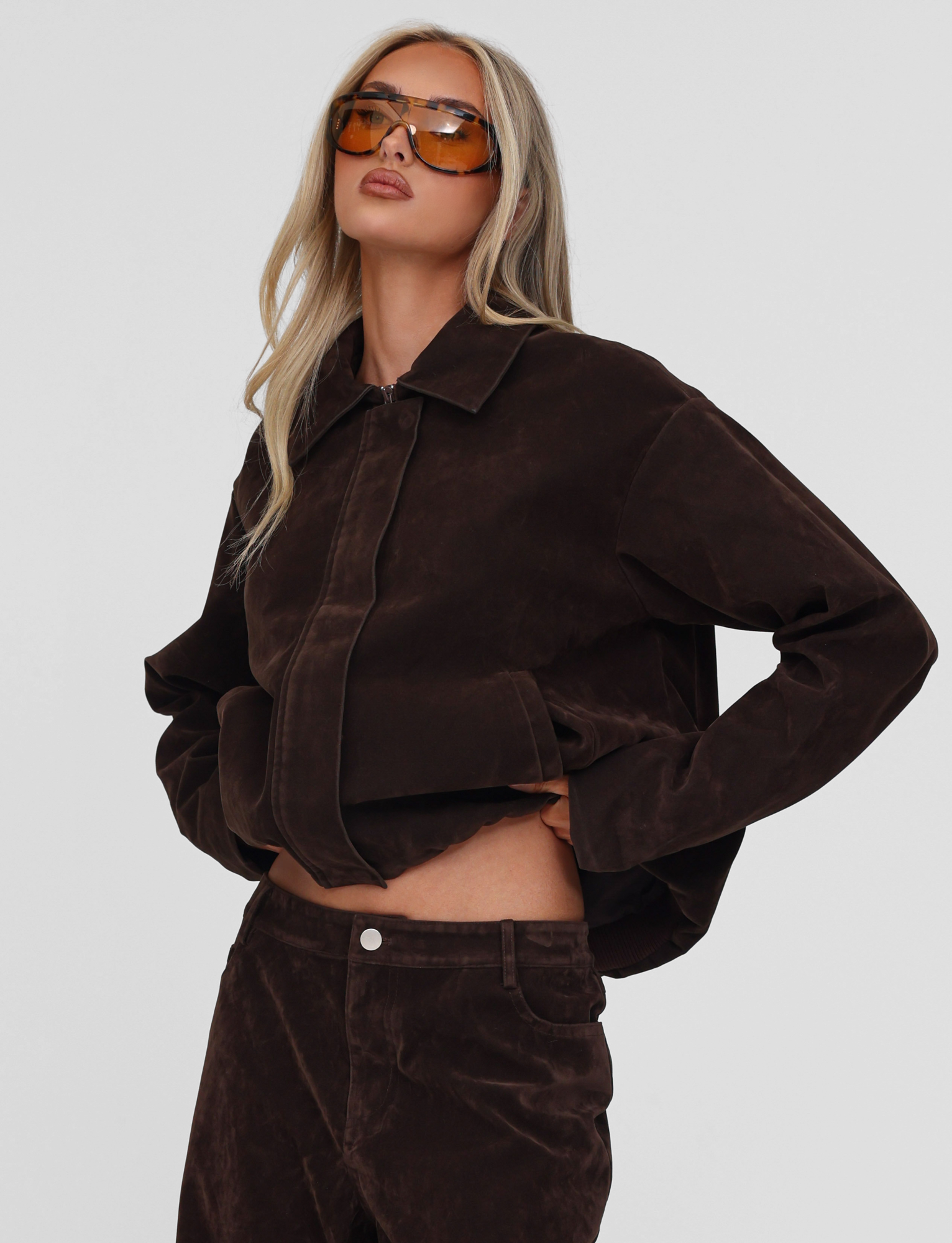 A woman with long blonde hair and large orange sunglasses poses confidently, wearing the TIGERMIST CAMELLIA BOMBER JACKET in chocolate suede with matching pants, against a plain white background.