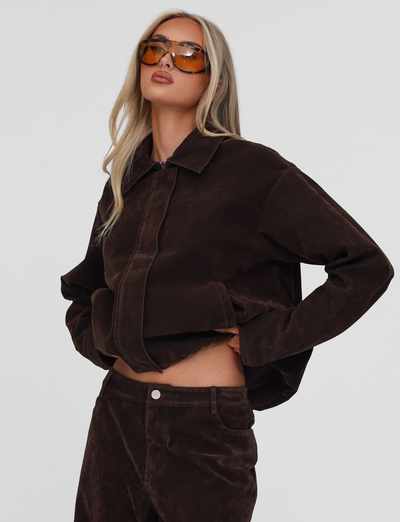 A woman with long blonde hair and large orange sunglasses poses confidently, wearing the TIGERMIST CAMELLIA BOMBER JACKET in chocolate suede with matching pants, against a plain white background.
