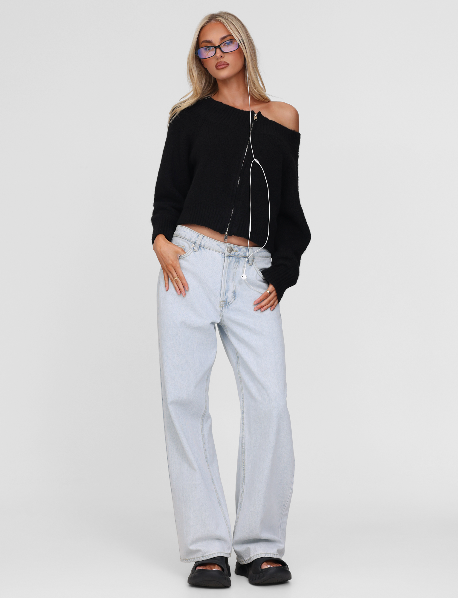 A woman stands against a plain background wearing the TIGERMIST LUNETTE JUMPER in black, paired with light-wash wide-leg jeans, black sandals, glasses, and earphones hanging around her neck.