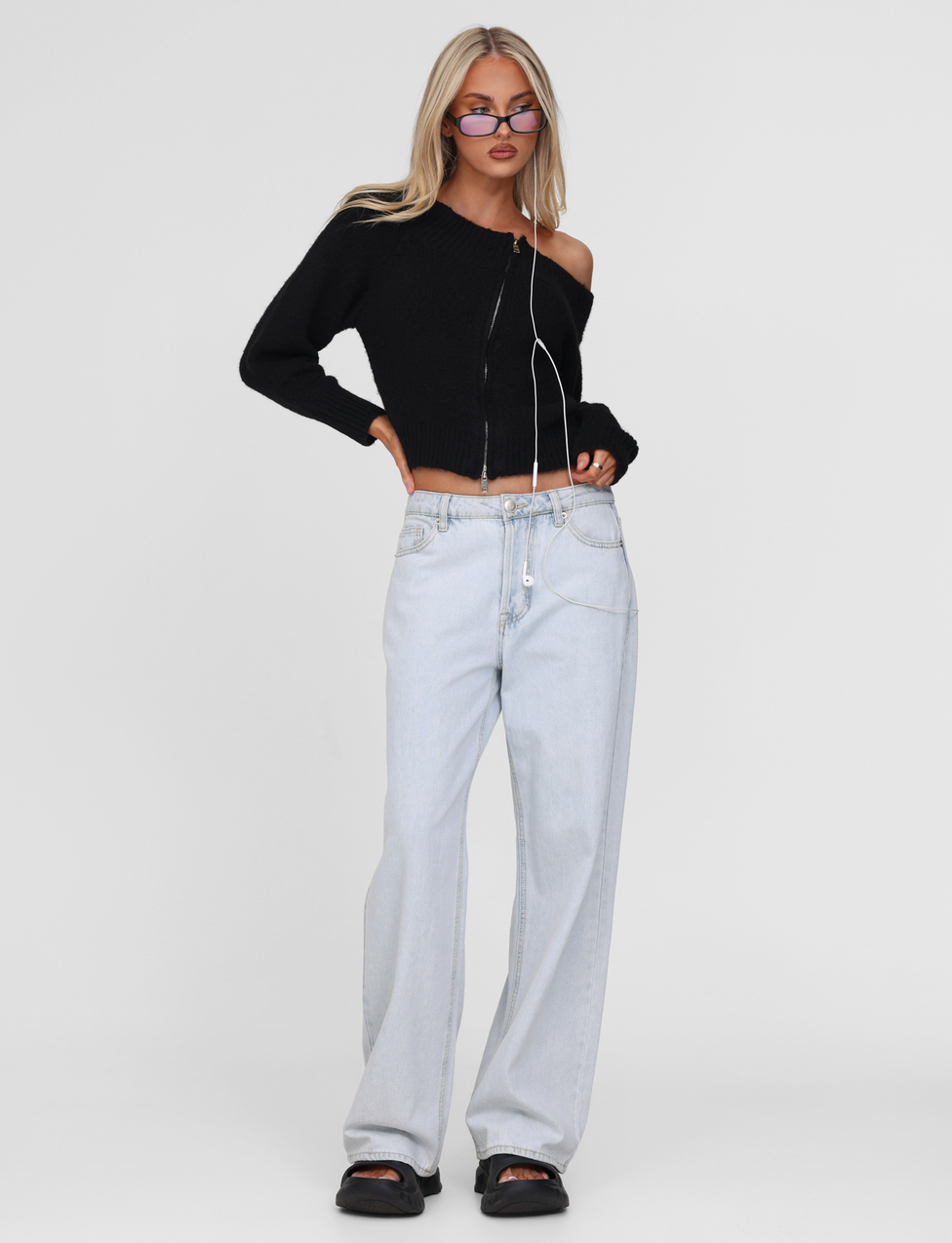 A woman with long blonde hair wears the TIGERMIST LUNETTE JUMPER in black, paired with light blue baggy jeans, black sandals, and purple sunglasses, standing against a plain white background.