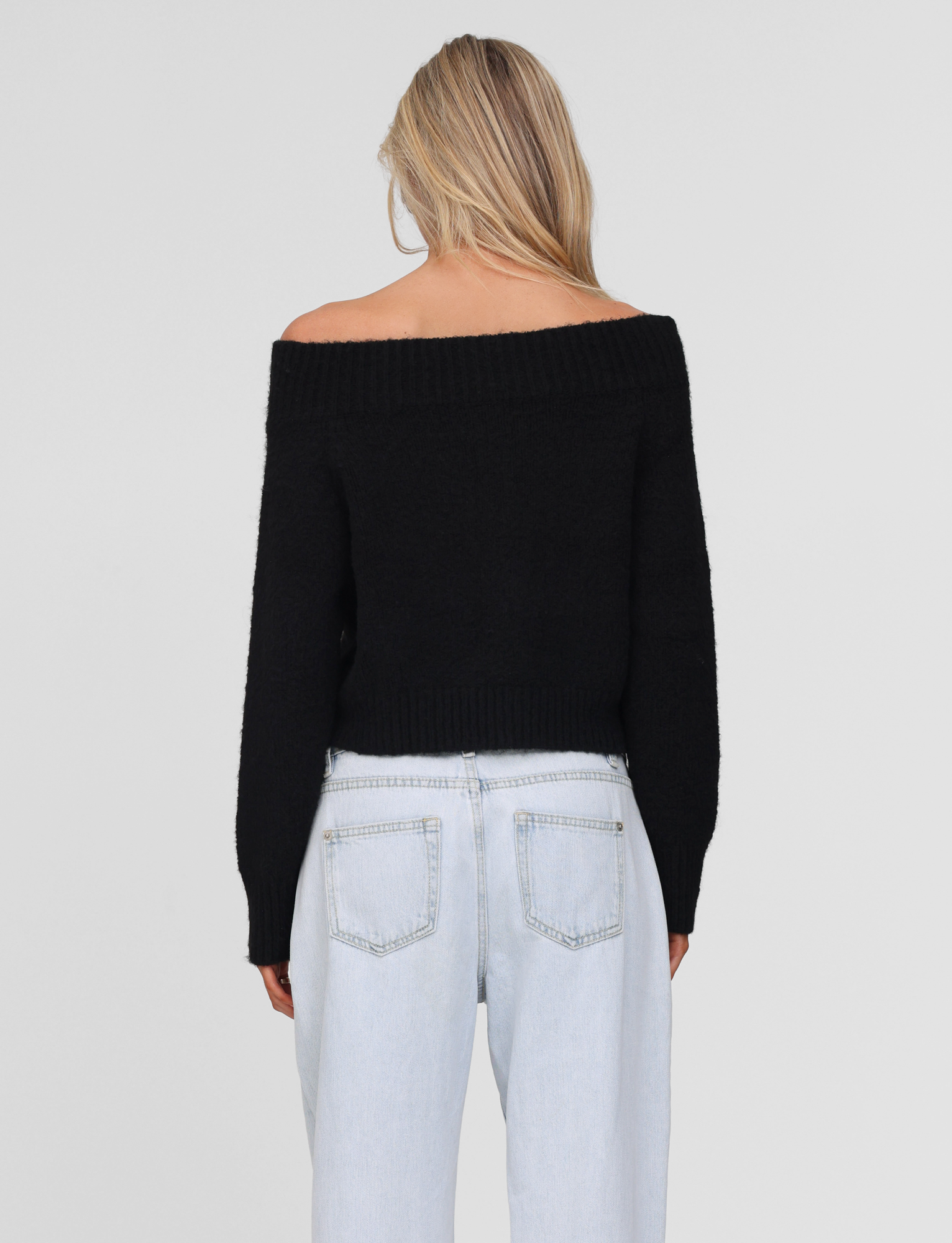 A woman with long blonde hair is seen from the back wearing the TIGERMIST LUNETTE JUMPER in black, paired with light blue high-waisted jeans against a plain light gray background.