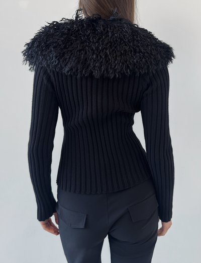 A person stands facing away, wearing the TIGERMIST BRISA JUMPER in black, which features a ribbed texture and a wide faux fur collar, paired with black pants with back pockets against a plain light background.