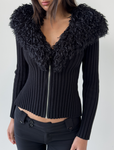 A woman wears the TIGERMIST BRISA JUMPER in black, a fitted ribbed jacket with a large faux fur collar and silver front zipper, paired with black pants. The jacket is zipped up; the photo is cropped at her head and mid-thigh.