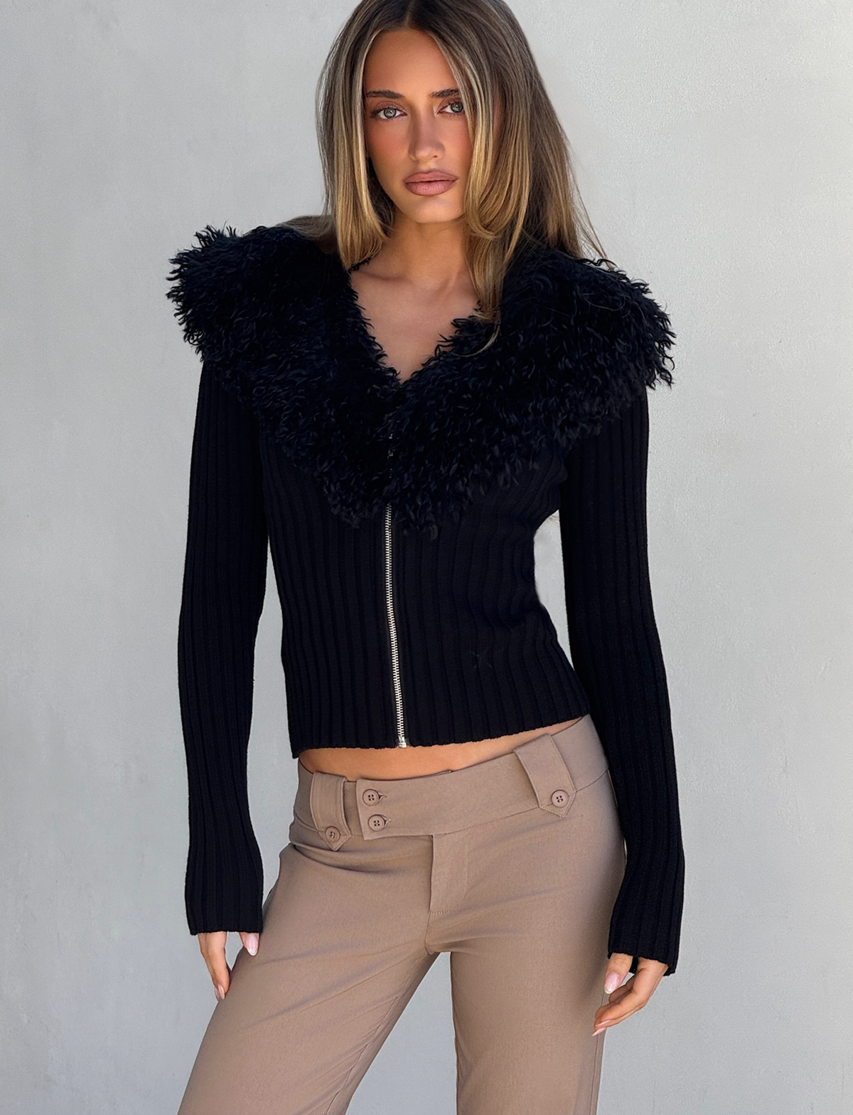 A woman models the TIGERMIST BRISA JUMPER in black—a ribbed cardigan with a faux fur collar and silver zipper—paired with beige pants, standing against a light gray background and looking at the camera with a neutral expression.