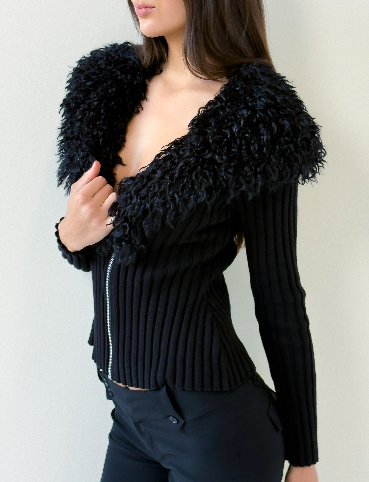 A woman wears the TIGERMIST BRISA JUMPER in black, featuring a ribbed design, large textured faux fur collar, and silver zipper, styled with black pants against a plain light background.
