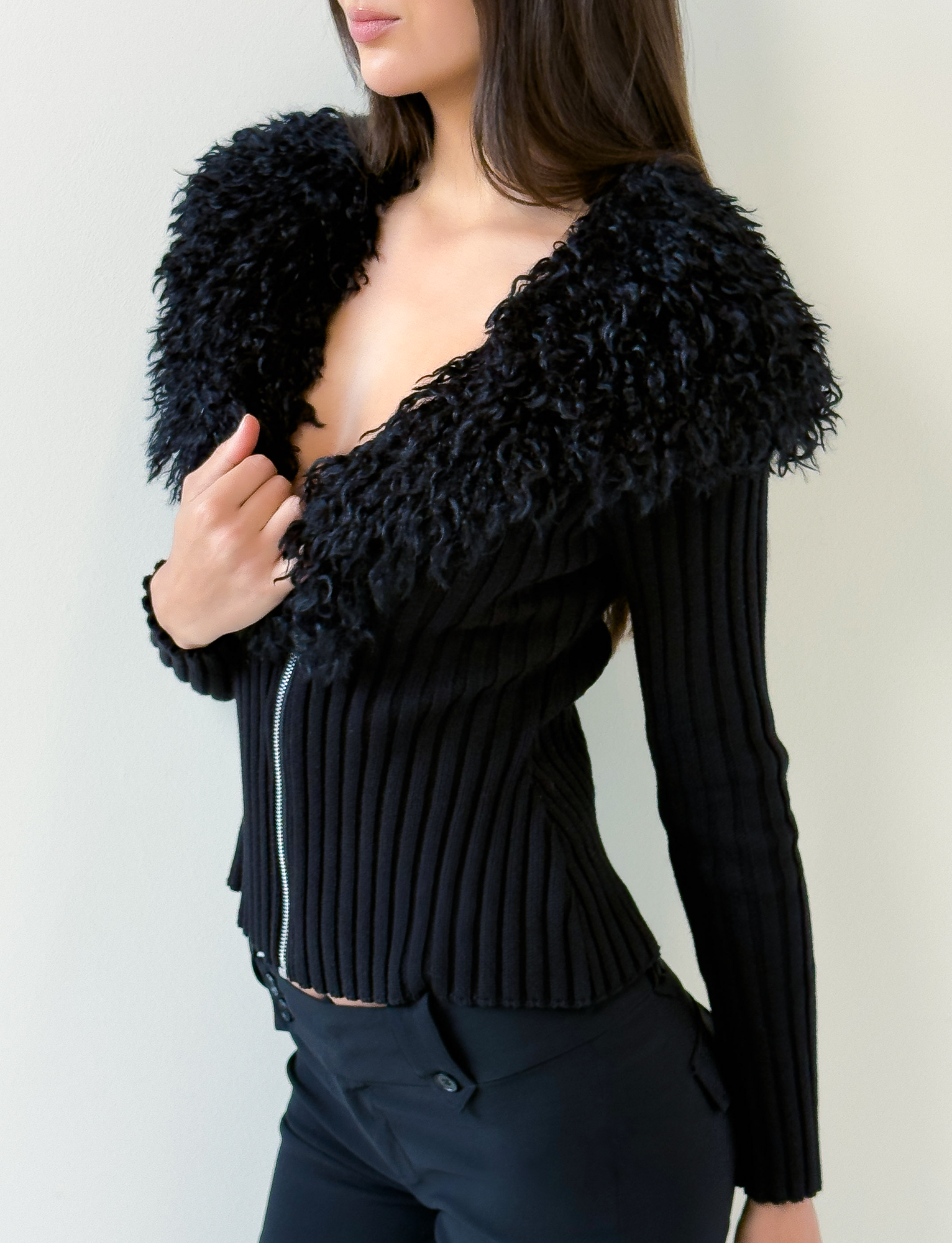A woman wears the TIGERMIST BRISA JUMPER in black, featuring a ribbed design, large textured faux fur collar, and silver zipper, styled with black pants against a plain light background.